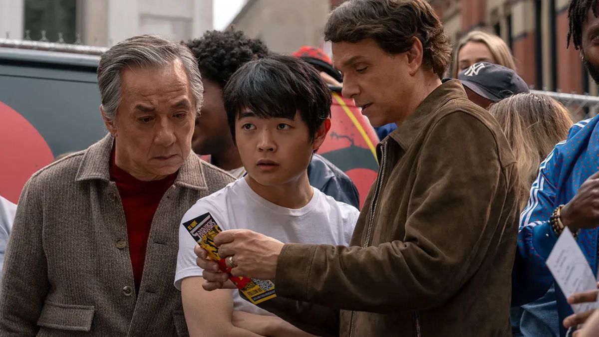 Three men in a crowd: an older Asian man, an Asian teen in a white t-shirt, and a man in a brown jacket showing something.