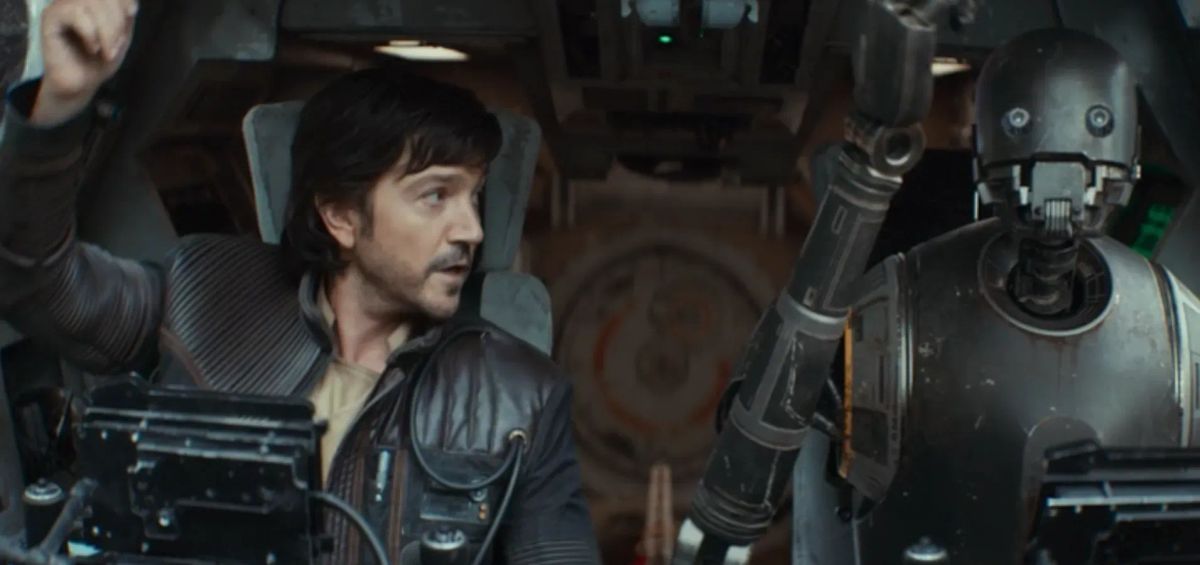 Man in leather jacket sits in a spacecraft cockpit next to a tall metallic droid, scene from Rogue One: A Star Wars Story.