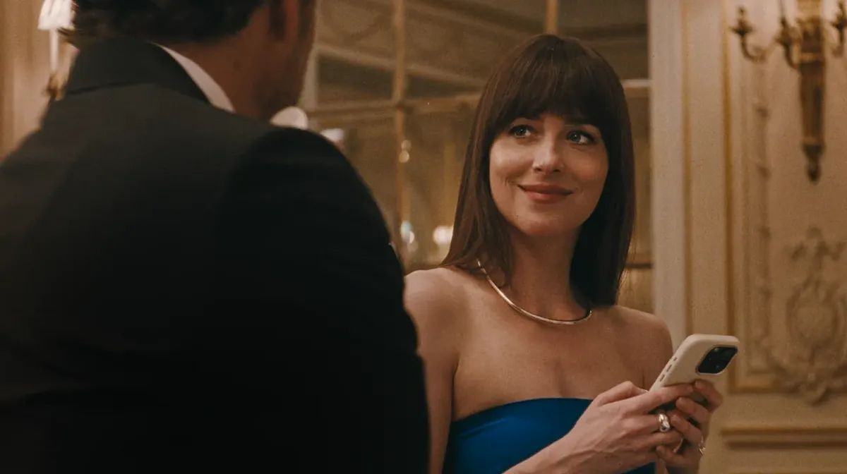 Woman in blue strapless dress with bangs holding a phone, smiling at a man in a dark suit in an elegant room with gold accents.