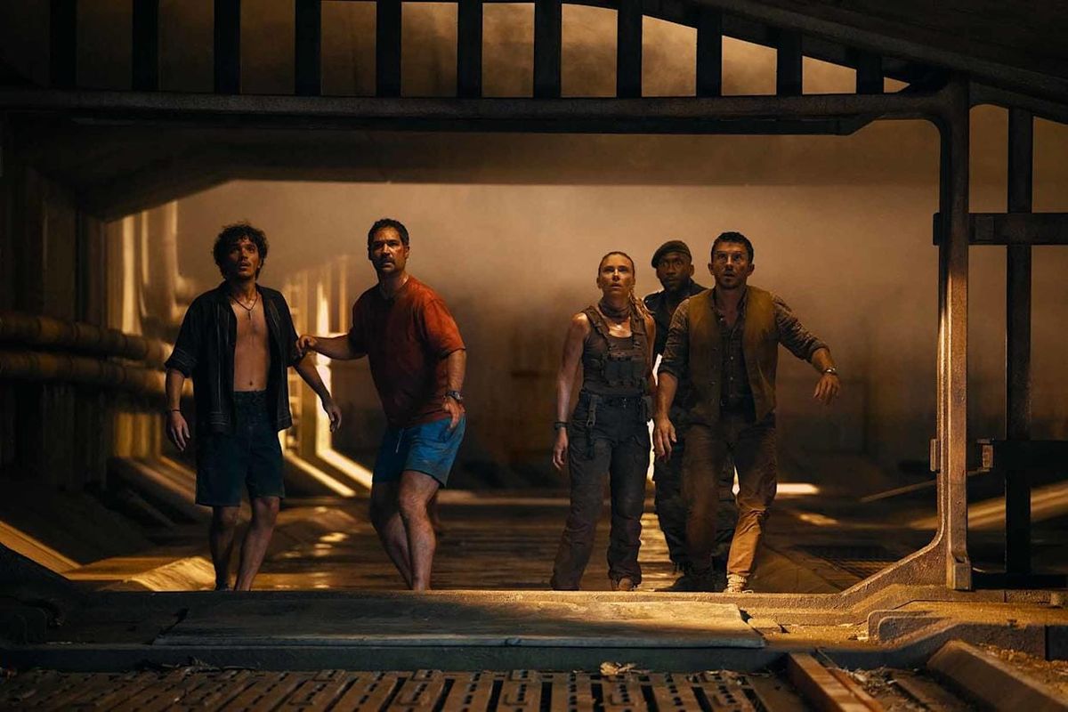 Five people walk cautiously through a dim, industrial corridor with pipes and metal grating, looking tense and alert.