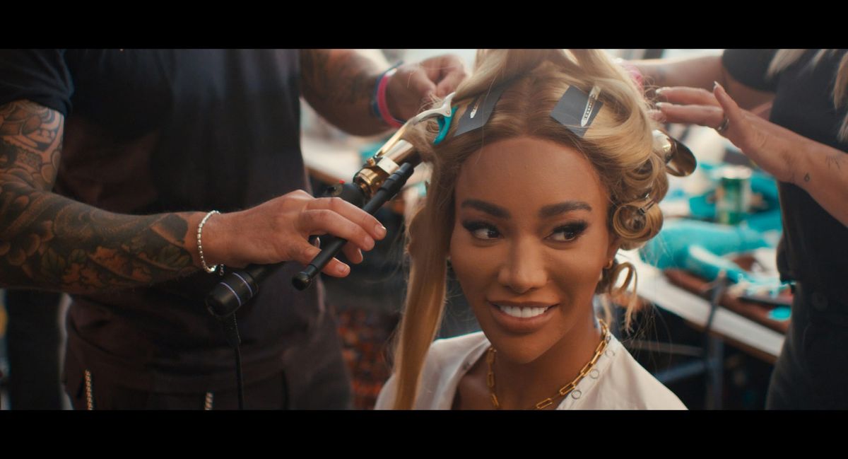 Woman with blonde hair getting styled with a curling iron by a tattooed hairstylist while another person clips sections.