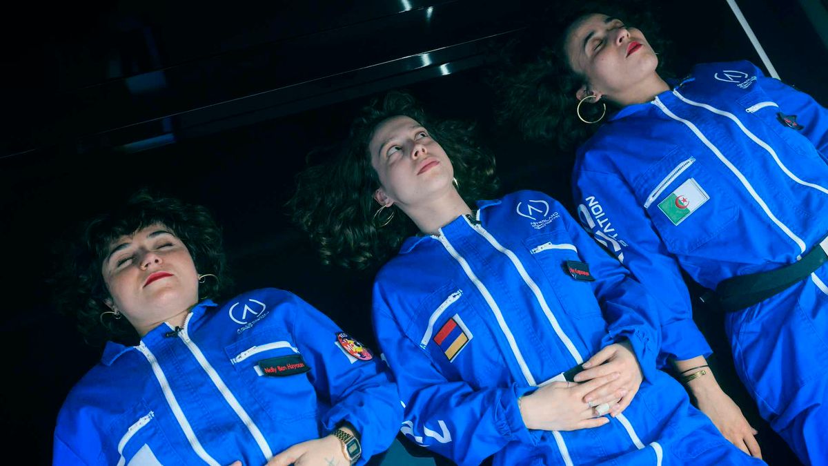 Three women in blue astronaut-style jumpsuits with patches and flags lie side by side with eyes closed against a dark background.