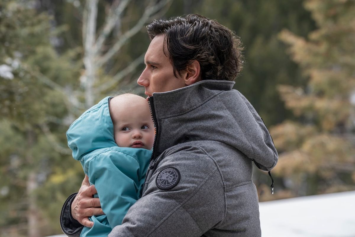 Man in gray Canada Goose jacket holding a baby in a light blue snowsuit outdoors with trees in the background.