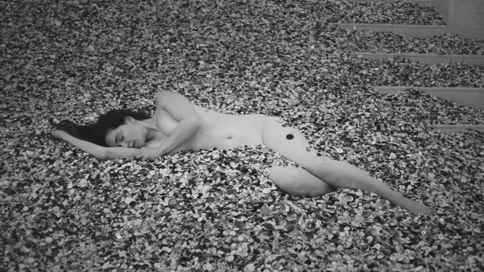 Black and white artistic photograph of a nude person lying on a thick bed of small flowers or petals near stone steps.