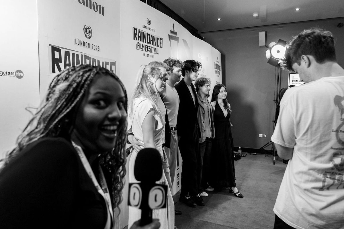 Black and white scene at Raindance Film Festival 2025 London with group posing for photos and a smiling woman holding a microphone.