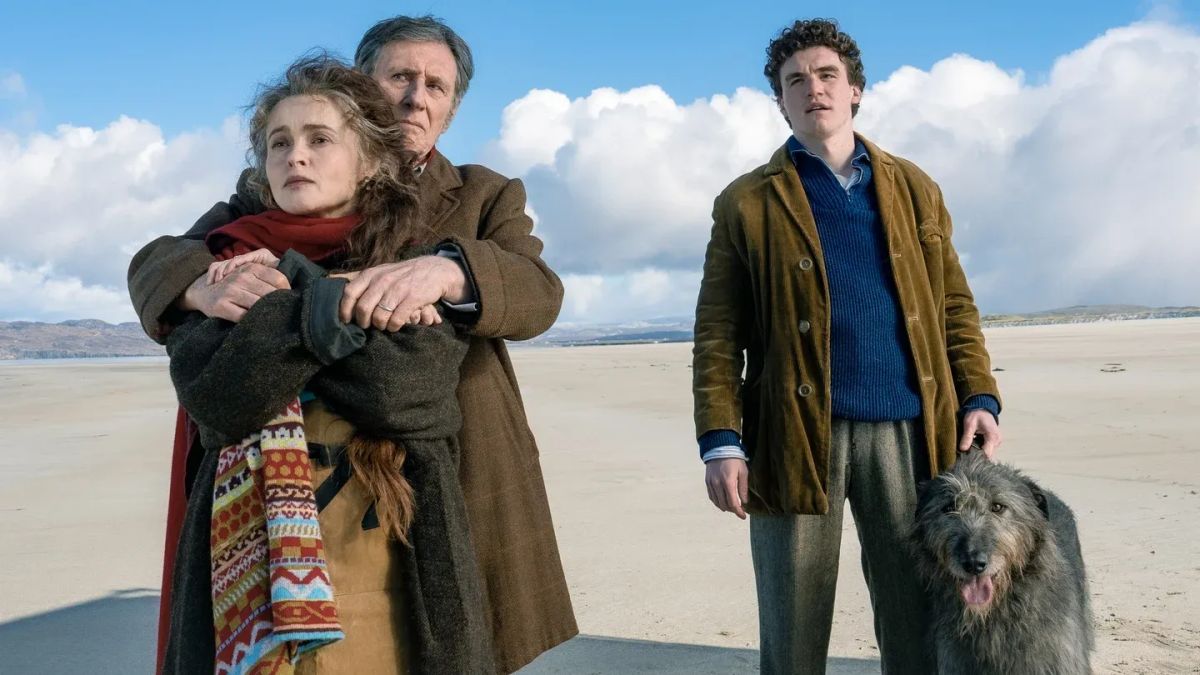 Three people on a windswept beach: a woman in a red scarf embraced by an older man in a brown coat, a young curly-haired man with a large shaggy dog.