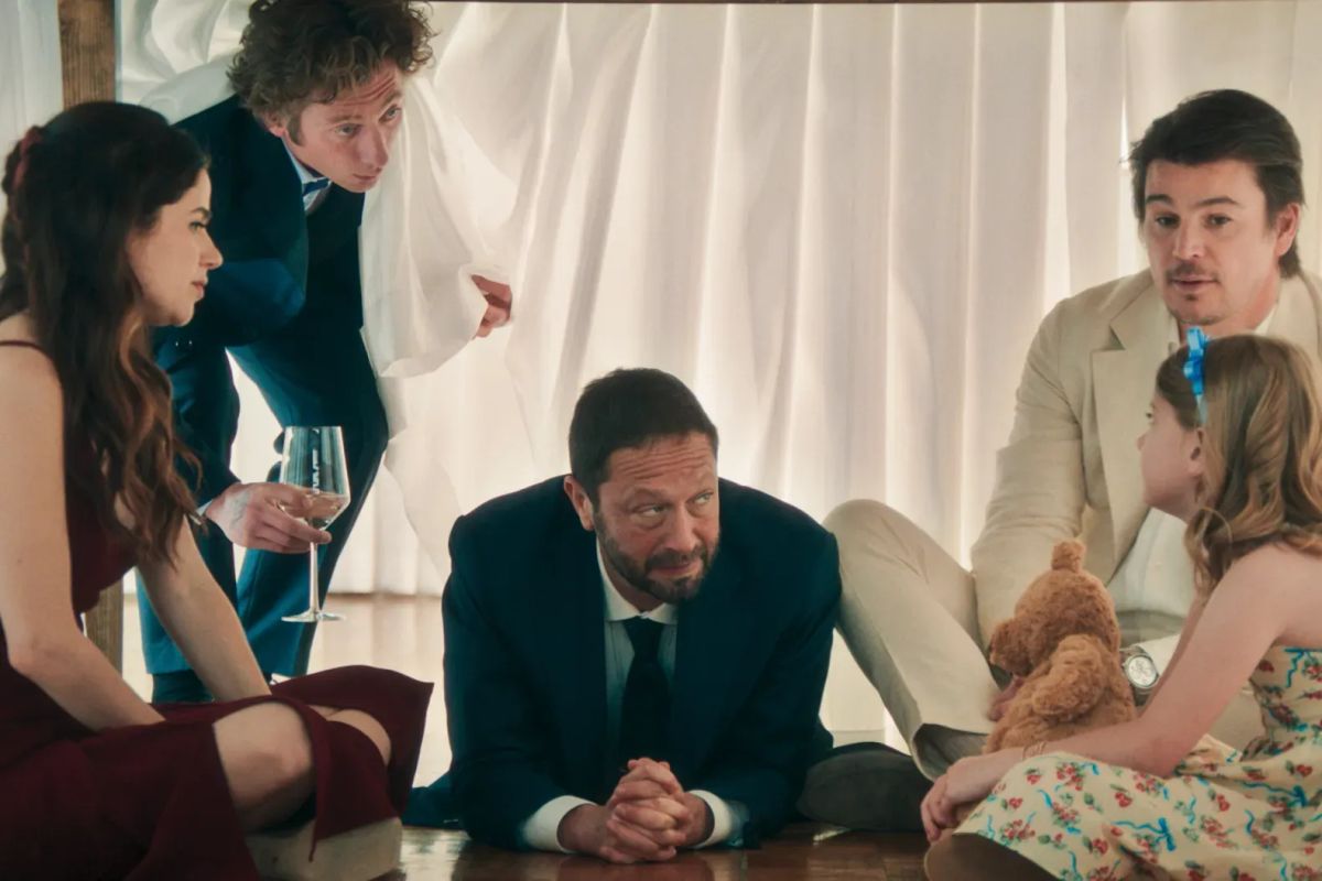 Five people huddle under a table with white draping; a man in a suit sits center, flanked by adults and a girl with a teddy bear.