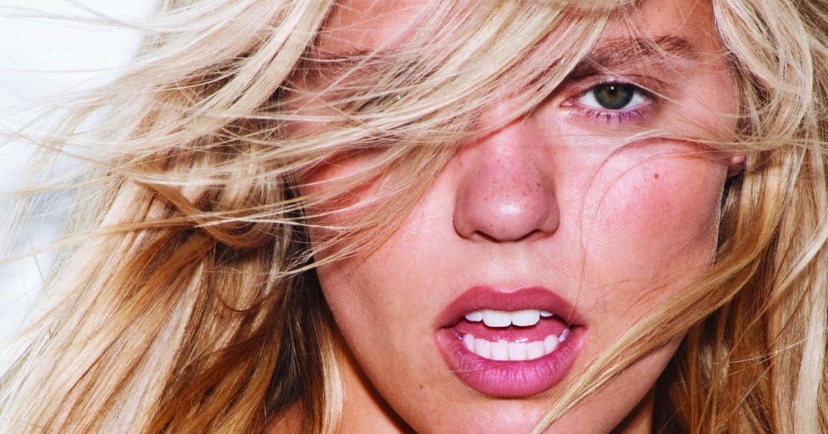 Close-up of a blonde woman with windswept hair, green eyes, pink lips, and sun-kissed skin gazing intensely at the camera.