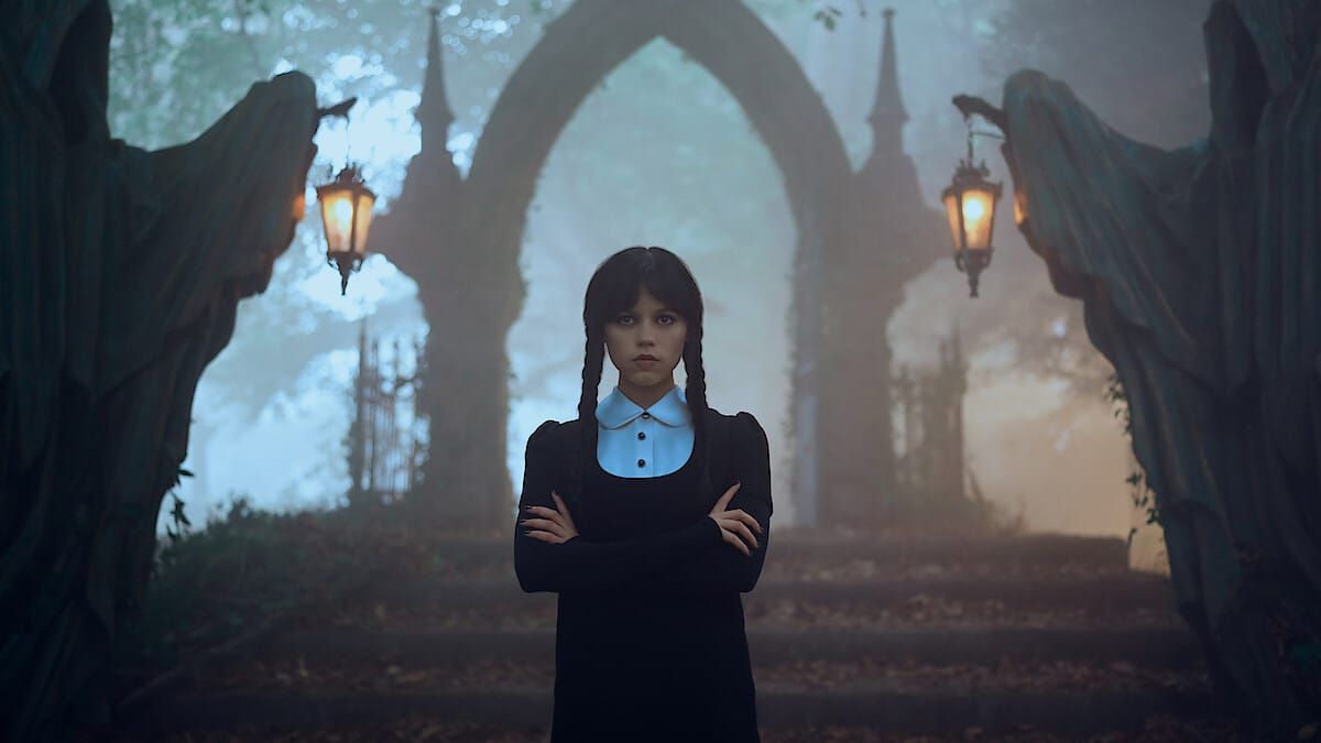 Young woman with dark braids in black dress and white collar stands with arms crossed before gothic archway with angel statues and lanterns in fog.
