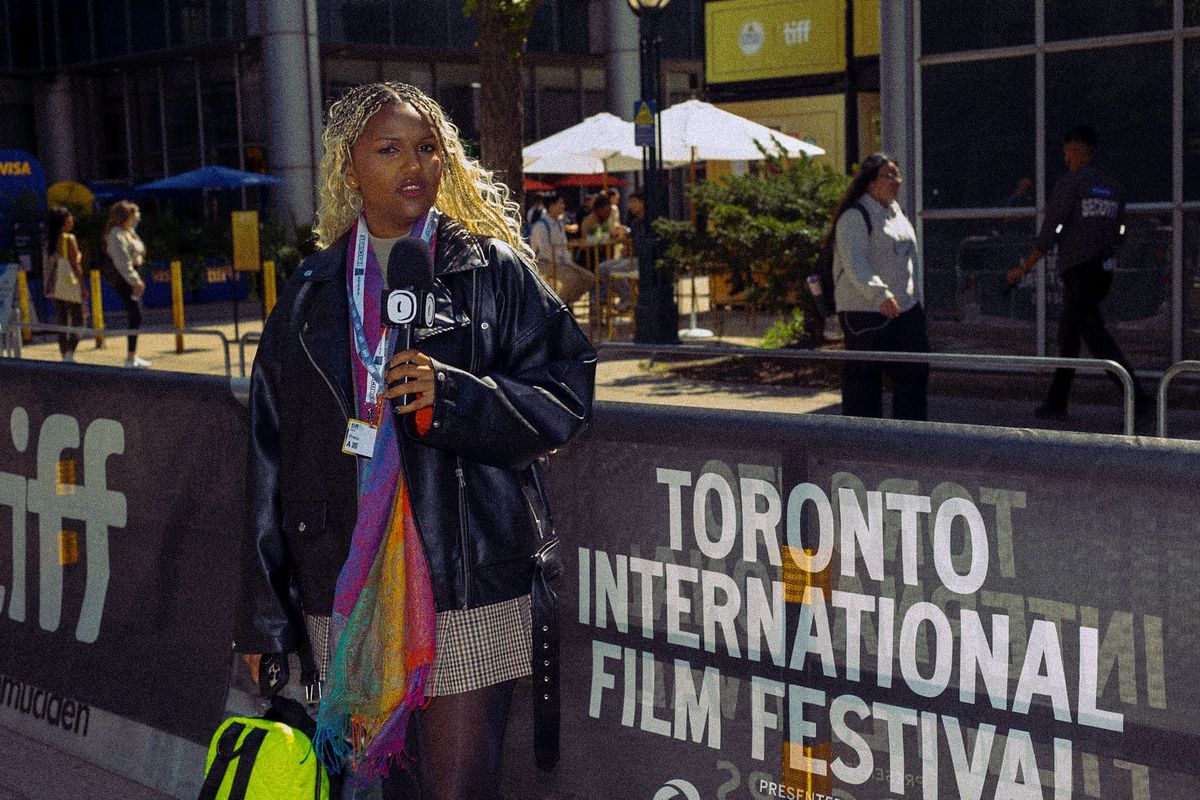 Reporter with blonde braids holds microphone outside Toronto International Film Festival, wearing leather jacket and lanyard.