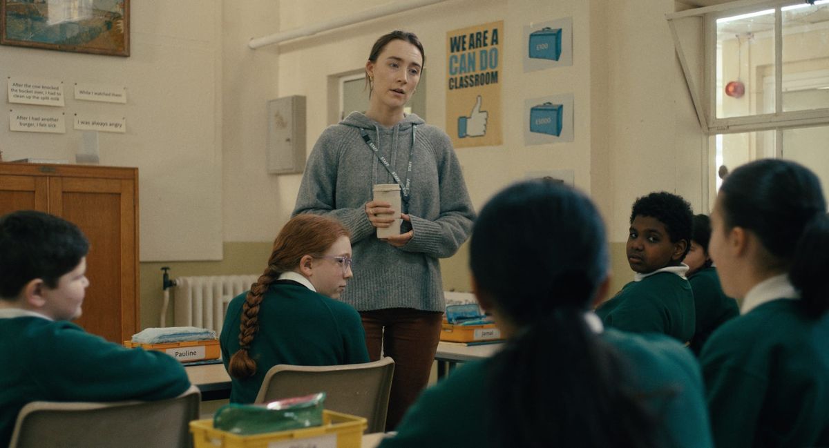 Teacher in gray hoodie holding a coffee cup addresses young students in green uniforms seated at desks in a classroom.