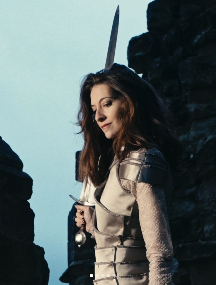 Woman with long brown hair in medieval-style armor holding a sword upright, standing among dark stone ruins against a pale sky.