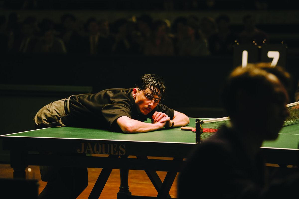 Man with glasses leaning exhausted on a green Jaques ping pong table, paddle nearby, crowd watching in dark arena background.