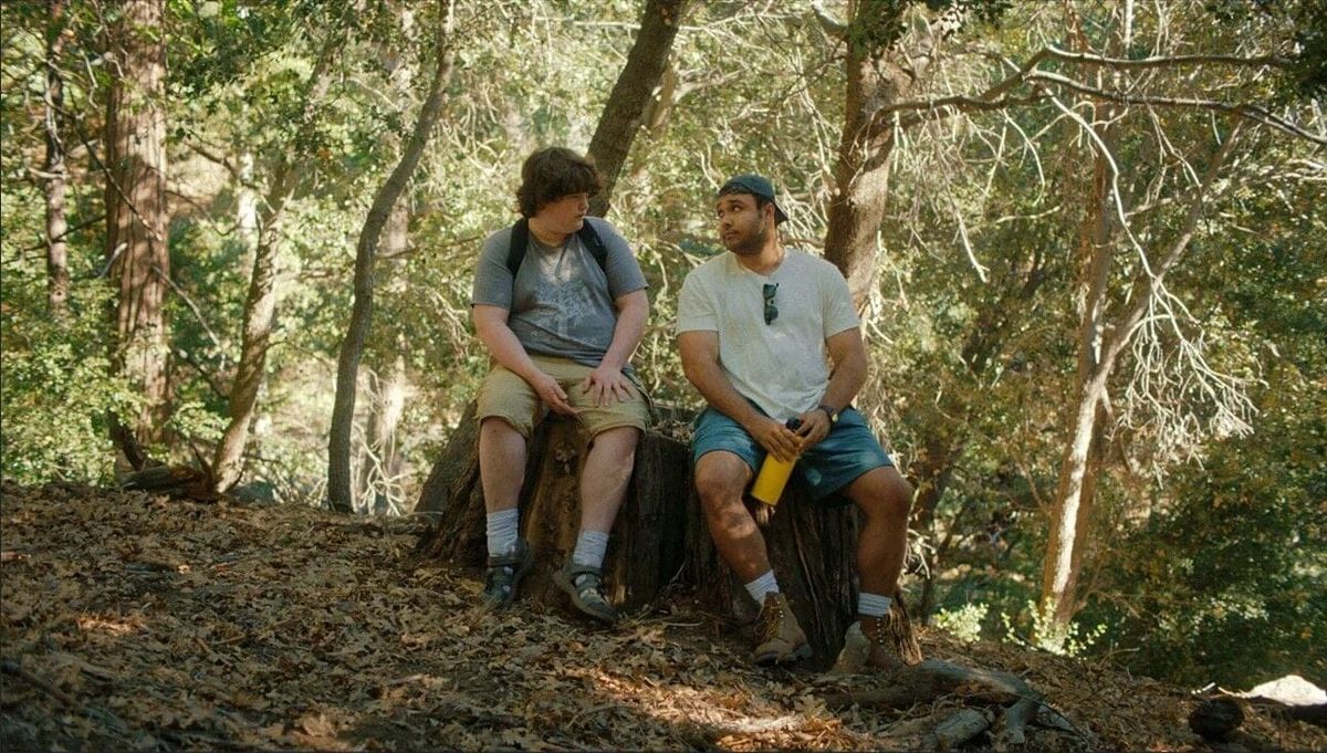 Two young men sitting on a tree stump in a wooded forest, one in a gray shirt and khaki shorts, the other in a white tee and blue shorts holding a yellow water bottle.