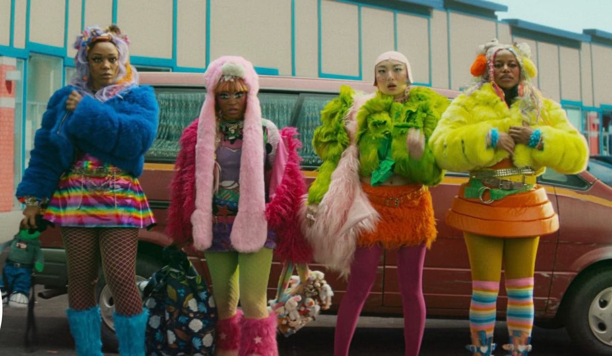 Four women in bold, colorful outfits with fuzzy coats and bright tights stand together in front of a red van in a parking lot.