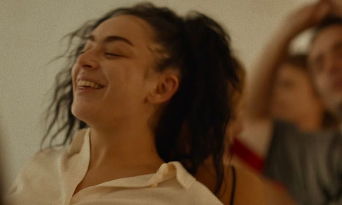 Young woman laughing joyfully with eyes closed and tousled dark hair, wearing a cream top, with a blurred person behind her.