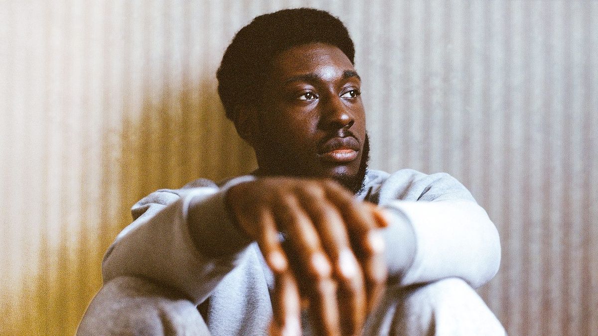 Young Black man in gray sweatshirt sitting with arms crossed over his knees, gazing intensely at camera against ridged wall.