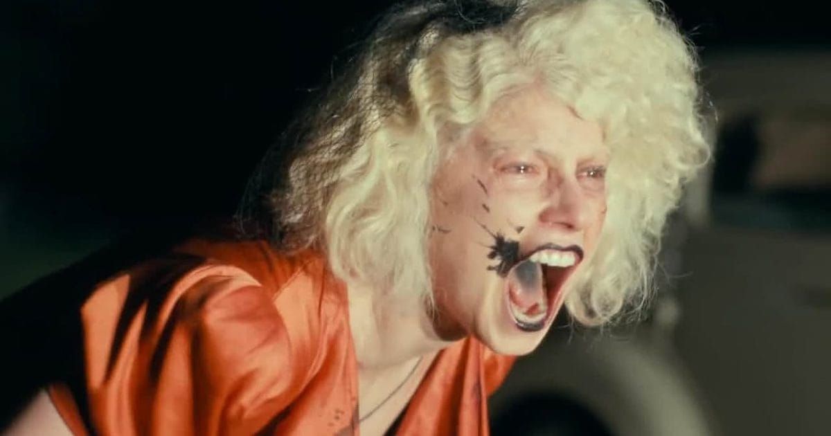 Person with wild blonde hair and smeared black face paint screaming, wearing an orange satin top, in a dark setting.