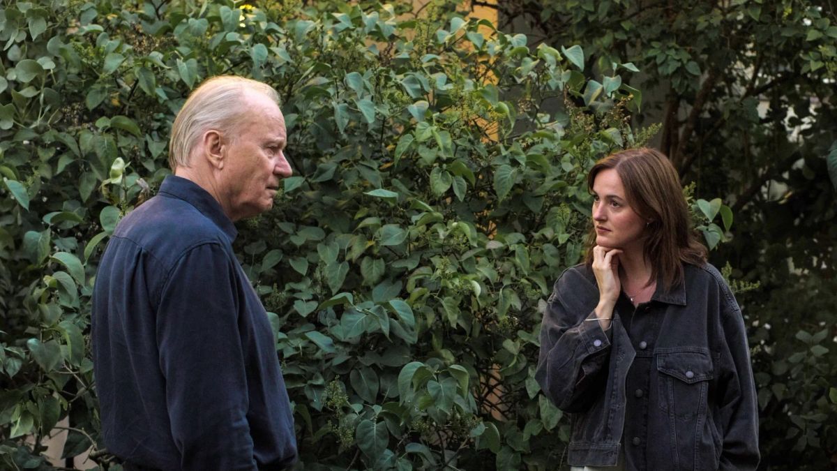 An older man in a navy shirt faces a younger woman in a dark denim jacket, talking outdoors near a lush green hedge.