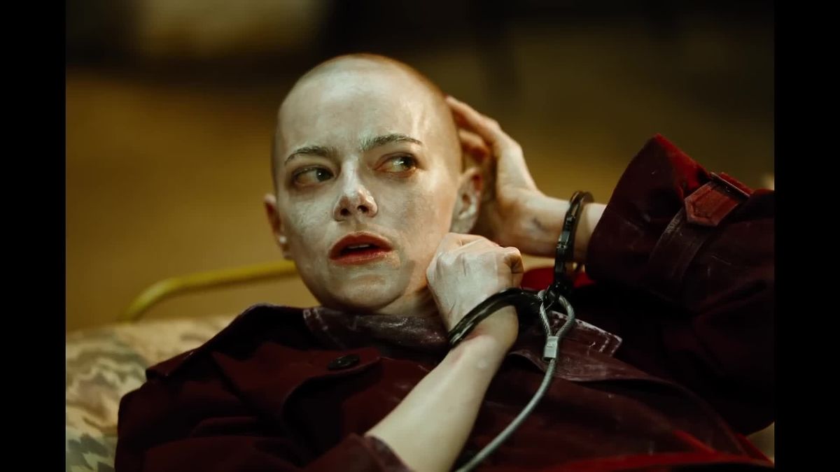 Bald person with pale white face makeup, wearing a dark red coat, handcuffed, lying down with a distressed expression.