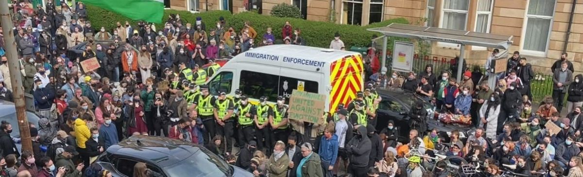 Crowd surrounds an Immigration Enforcement van while police in high-vis jackets form a cordon around it on a city street.