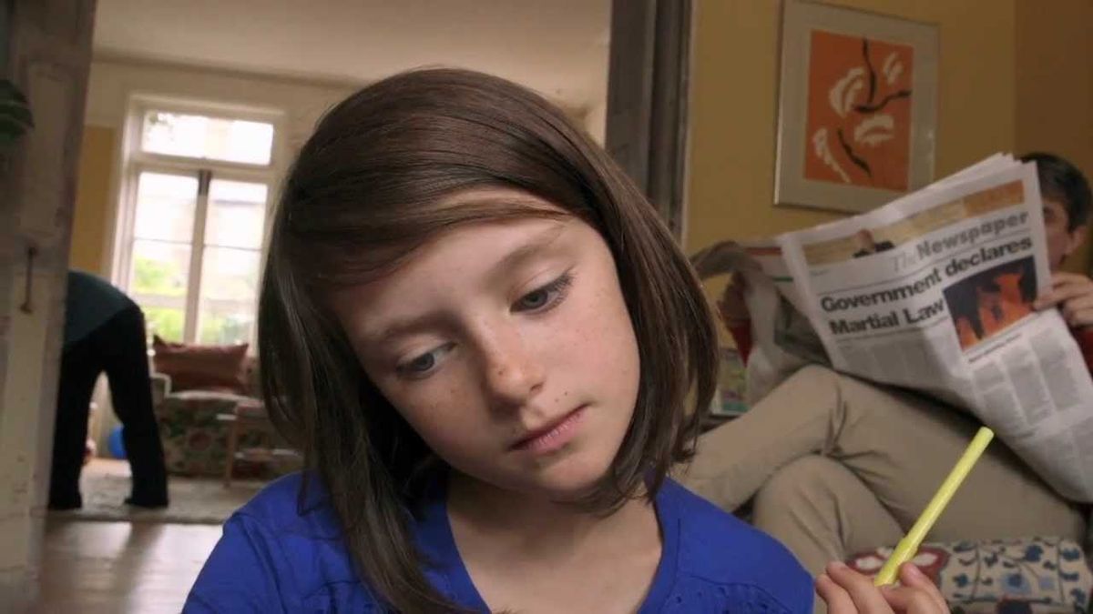 Young girl with brown bob hair in blue shirt holds yellow pencil; behind her, person reads newspaper headlined "Government declares Martial Law"