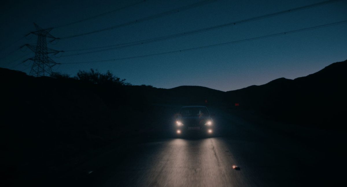 A car travels down a road at twilight with its headlights on, darkness surrounding it.