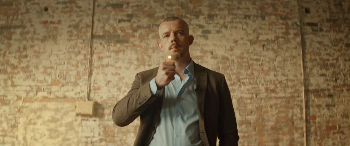 Man with shaved head in brown blazer and light blue shirt holding a lit match, standing before an exposed brick wall.