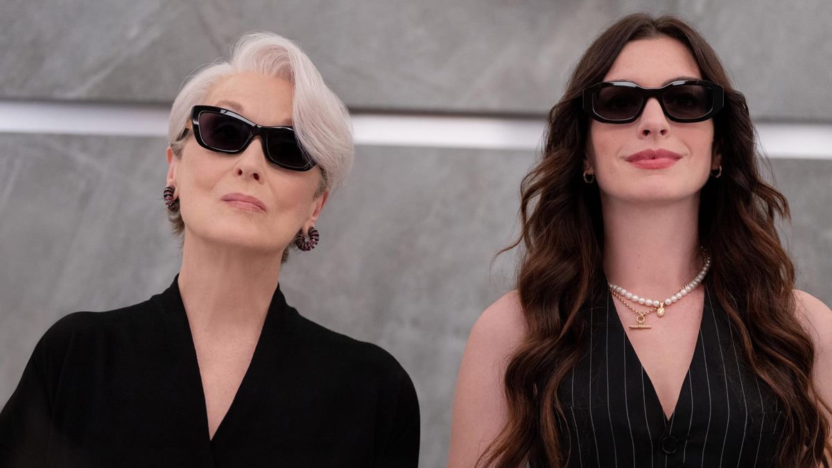 Two women in black outfits and dark sunglasses seated together; one older with short white hair, one younger with long brown hair and a pearl necklace.