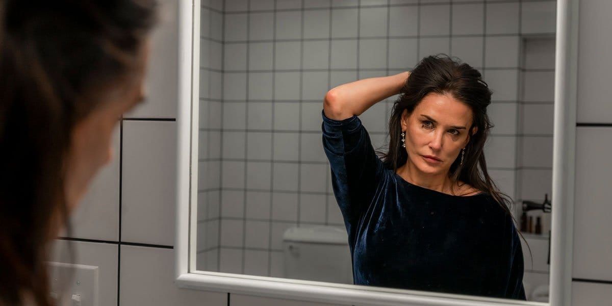 Woman in dark blue velvet top looking at her reflection in a bathroom mirror while running her hand through her dark hair.