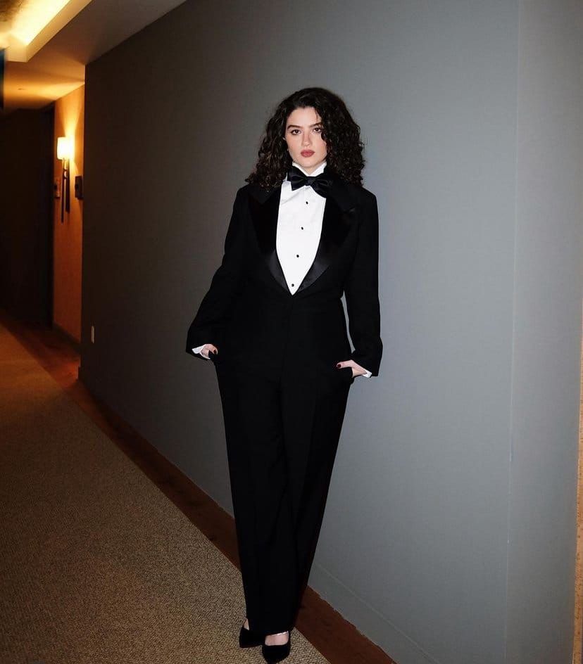 Young woman with curly dark hair wearing a black tuxedo with bow tie, standing in a hotel hallway with hands in pockets.