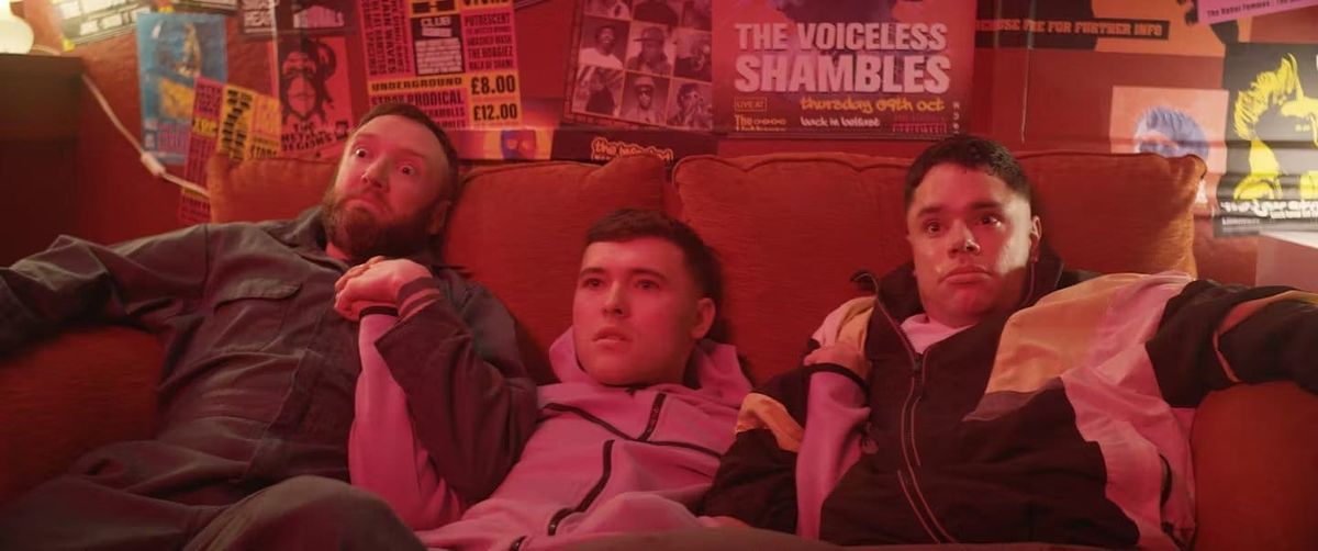 Three young men sitting closely together on a red couch surrounded by music gig posters in a dimly lit, red-toned room.