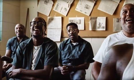 Four men sitting together laughing in a room with papers pinned to a bulletin board behind them, wearing casual uniforms.