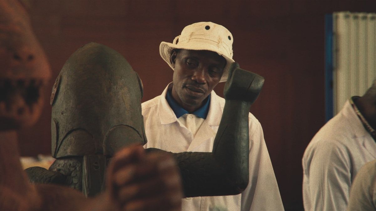 Man in white bucket hat and shirt examines a large bronze sculpture in a warmly lit workshop or studio.