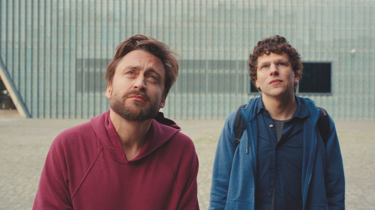 Two men standing outdoors looking upward; one in a red hoodie with brown hair and beard, the other in a blue jacket with curly hair and glasses.