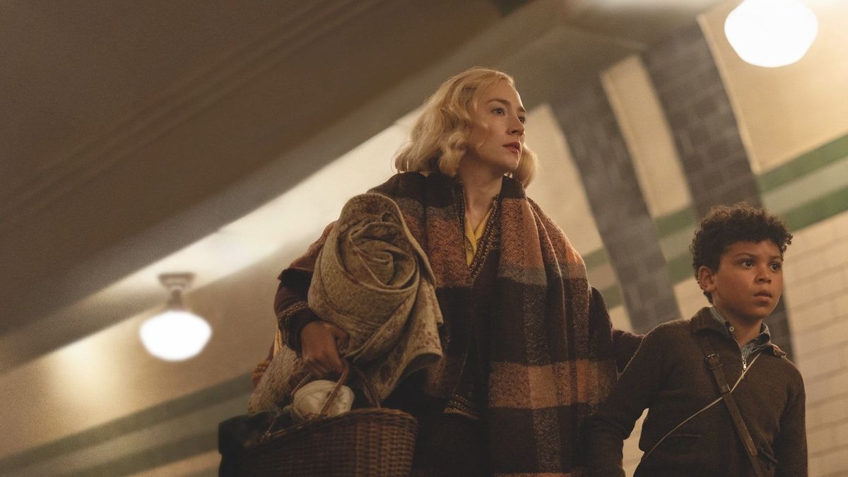 Blonde woman in plaid shawl carrying a rolled blanket and basket stands beside a young curly-haired boy in a dimly lit station.