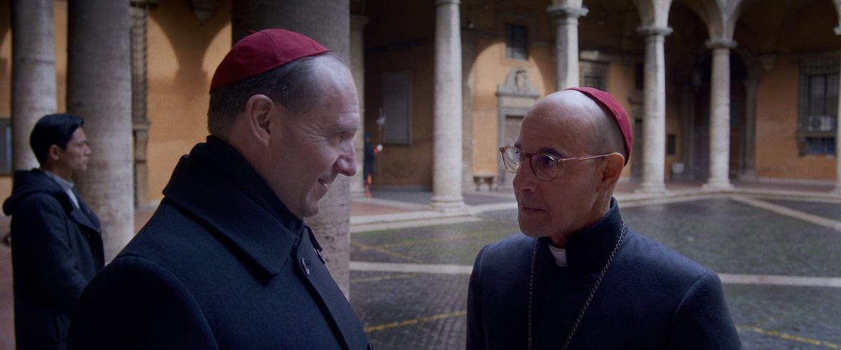 Two Catholic clergy in dark cassocks and red zucchetto caps converse in an Italian courtyard with stone columns and arches.