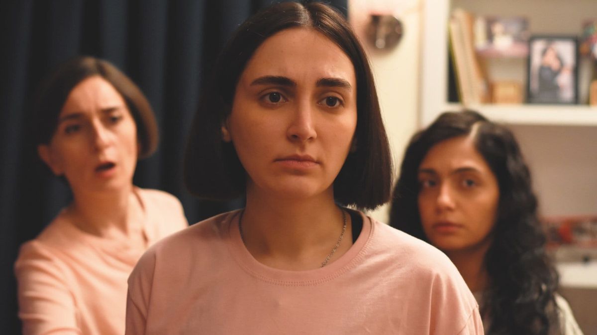 Three young women with concerned expressions in a room; center woman in pink top looks directly at camera, two others blurred behind.