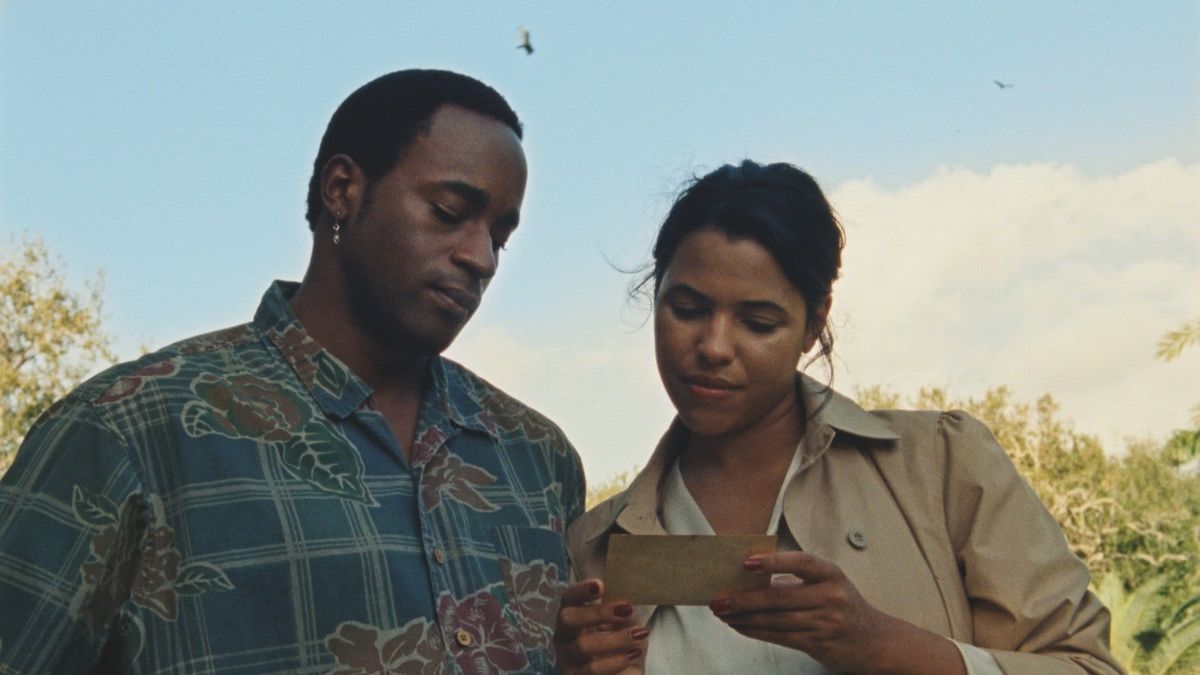 Two people standing outdoors looking at a small card or photograph the woman is holding, with blue sky and greenery behind them.