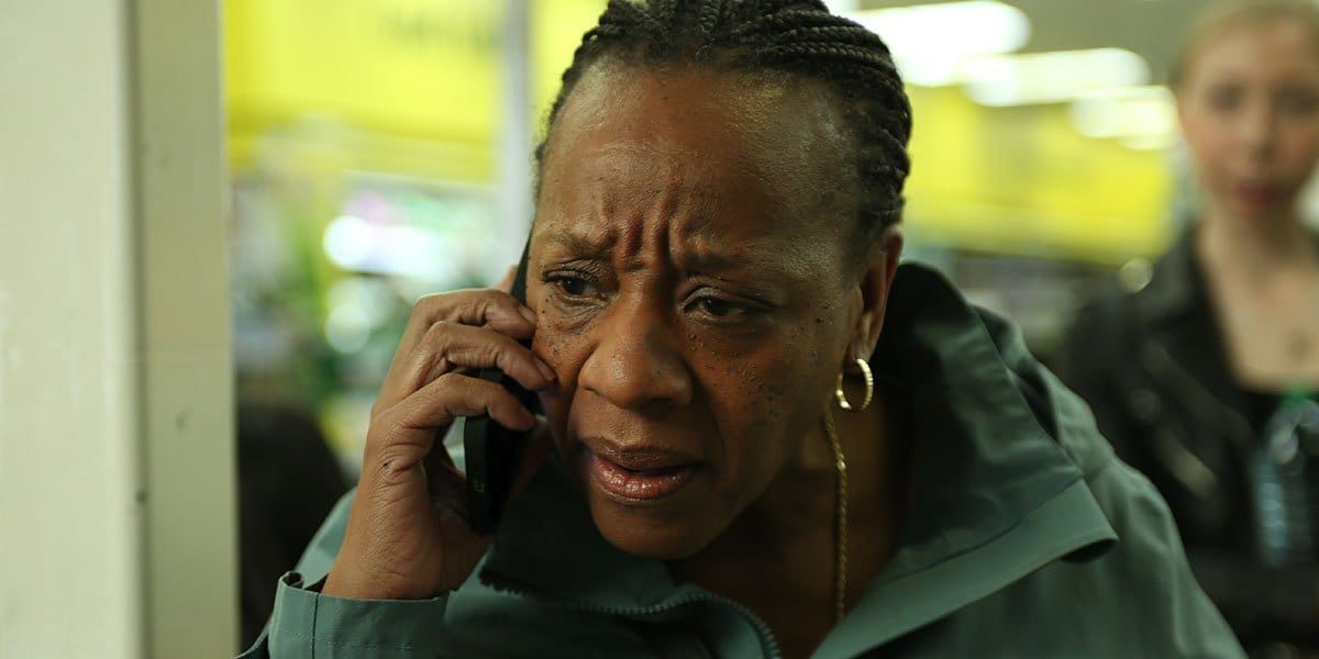 Middle-aged Black woman with braided hair talking on a cellphone, looking concerned, wearing a dark green jacket and gold hoop earrings.
