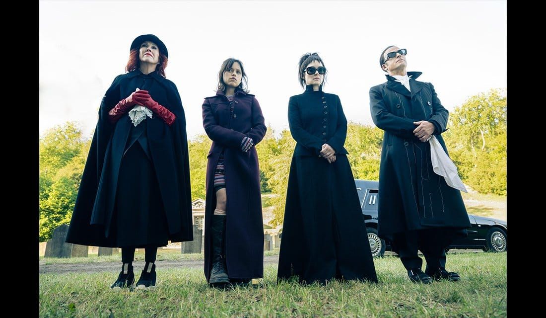 Four people in dark gothic clothing stand in a graveyard, wearing long black coats and capes, with trees and a car behind.