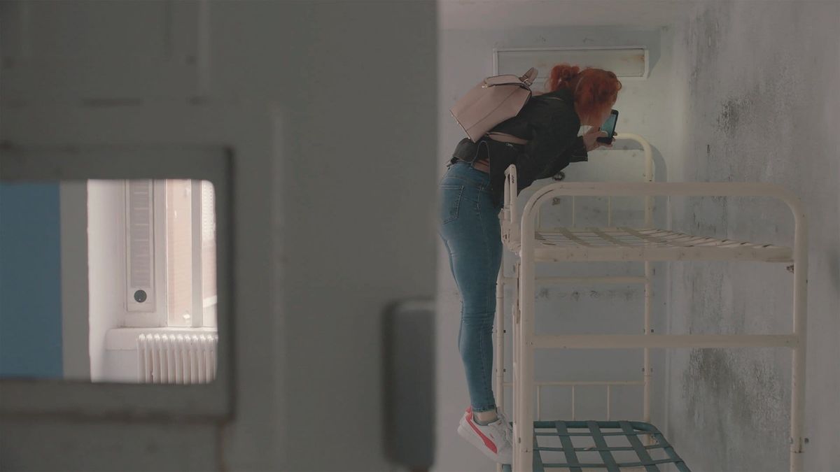 Red-haired woman in jeans and black jacket climbs a bunk bed frame while holding a phone, viewed through a prison cell door.