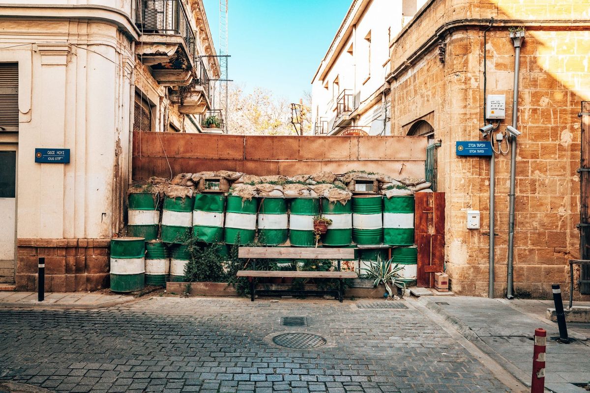 Green-and-white oil drum barricade topped with sandbags blocking a street in Nicosia's UN Buffer Zone, Cyprus, with street signs and a bench.
