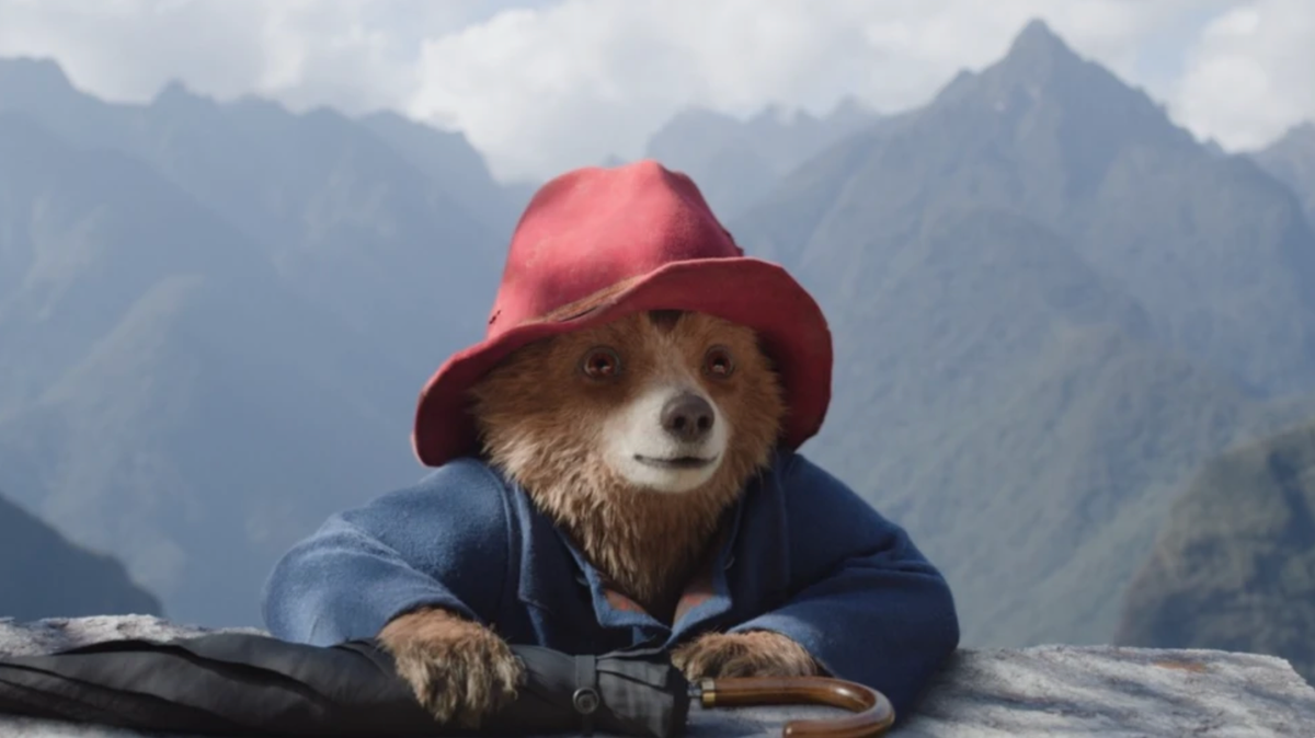 CGI Paddington Bear wearing his red hat and blue duffle coat, leaning on a ledge with mountains in the background.