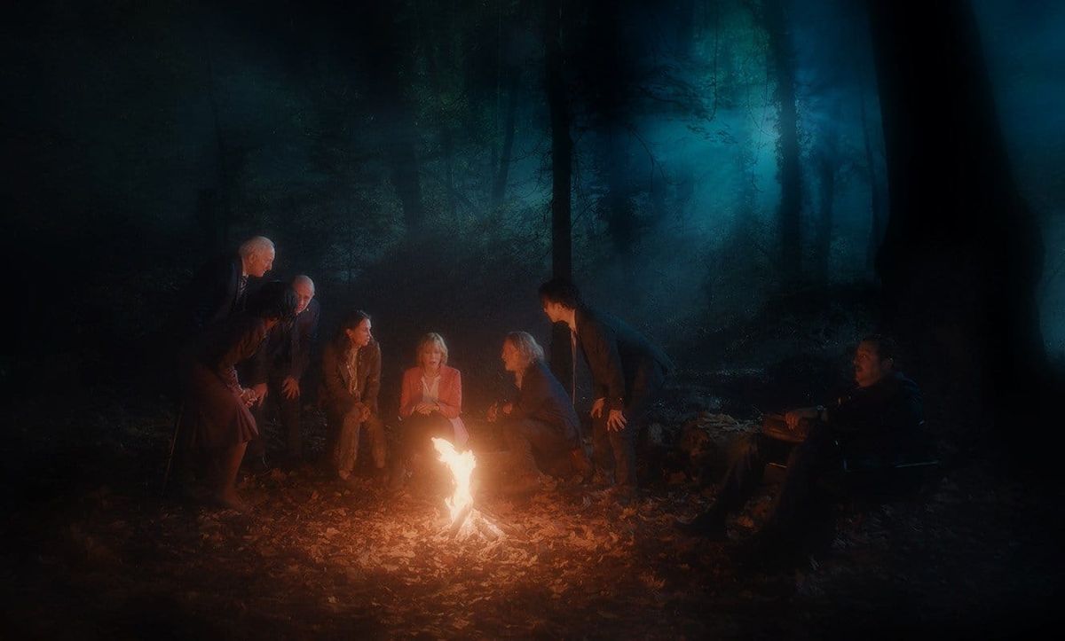 Group of eight people gathered around a small campfire in a dark, misty forest at night, illuminated by warm firelight.