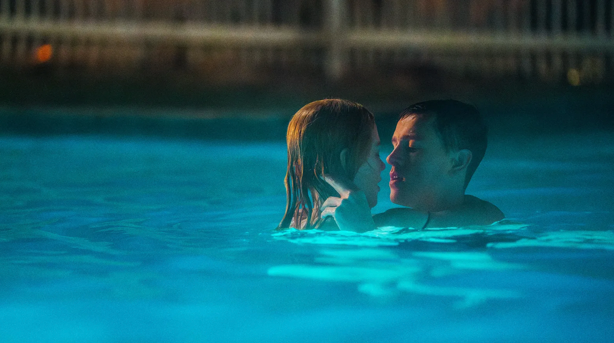 Two people embracing closely in a blue-lit swimming pool at night, faces near each other in an intimate moment.