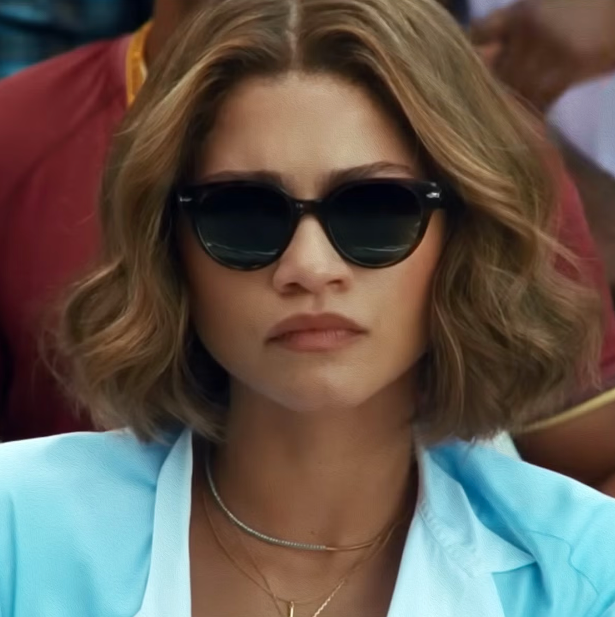 Woman with wavy shoulder-length blonde hair wearing dark sunglasses, gold layered necklaces, and a light blue blazer.