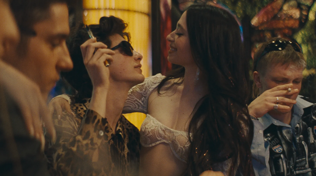 Young people at a party; a man in a leopard-print shirt adjusts his sunglasses while a woman in a white lace top laughs beside him.