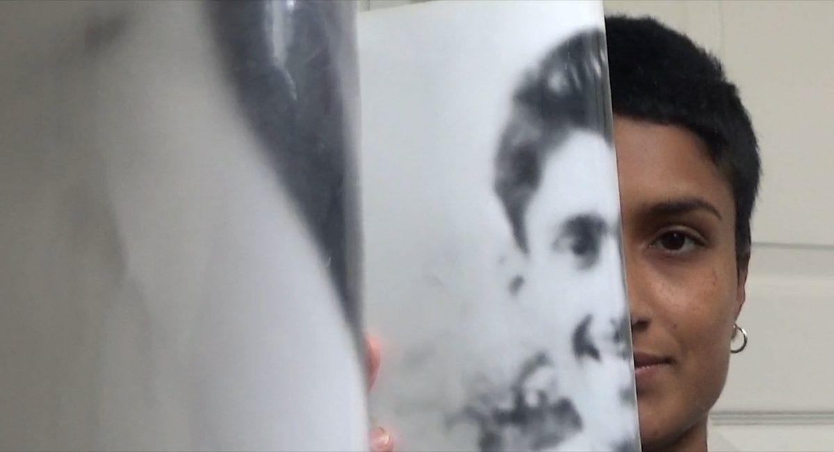 Person with short dark hair and hoop earring peeks from behind a black-and-white printed photograph of someone's face.