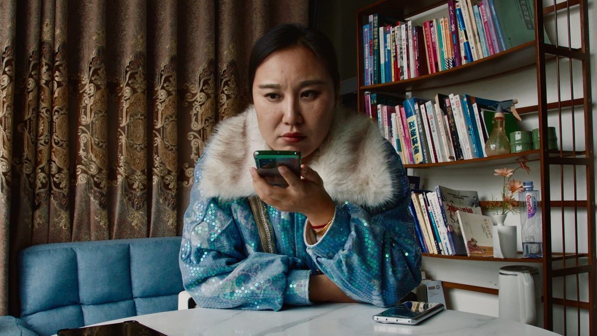Woman in blue sequined jacket with white fur collar holds phone near her face, sitting at white desk with bookshelf behind.